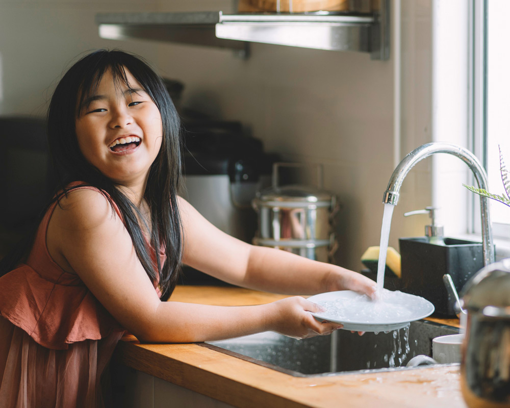 Girl laughs as she rinses her plate at the kitchen sink